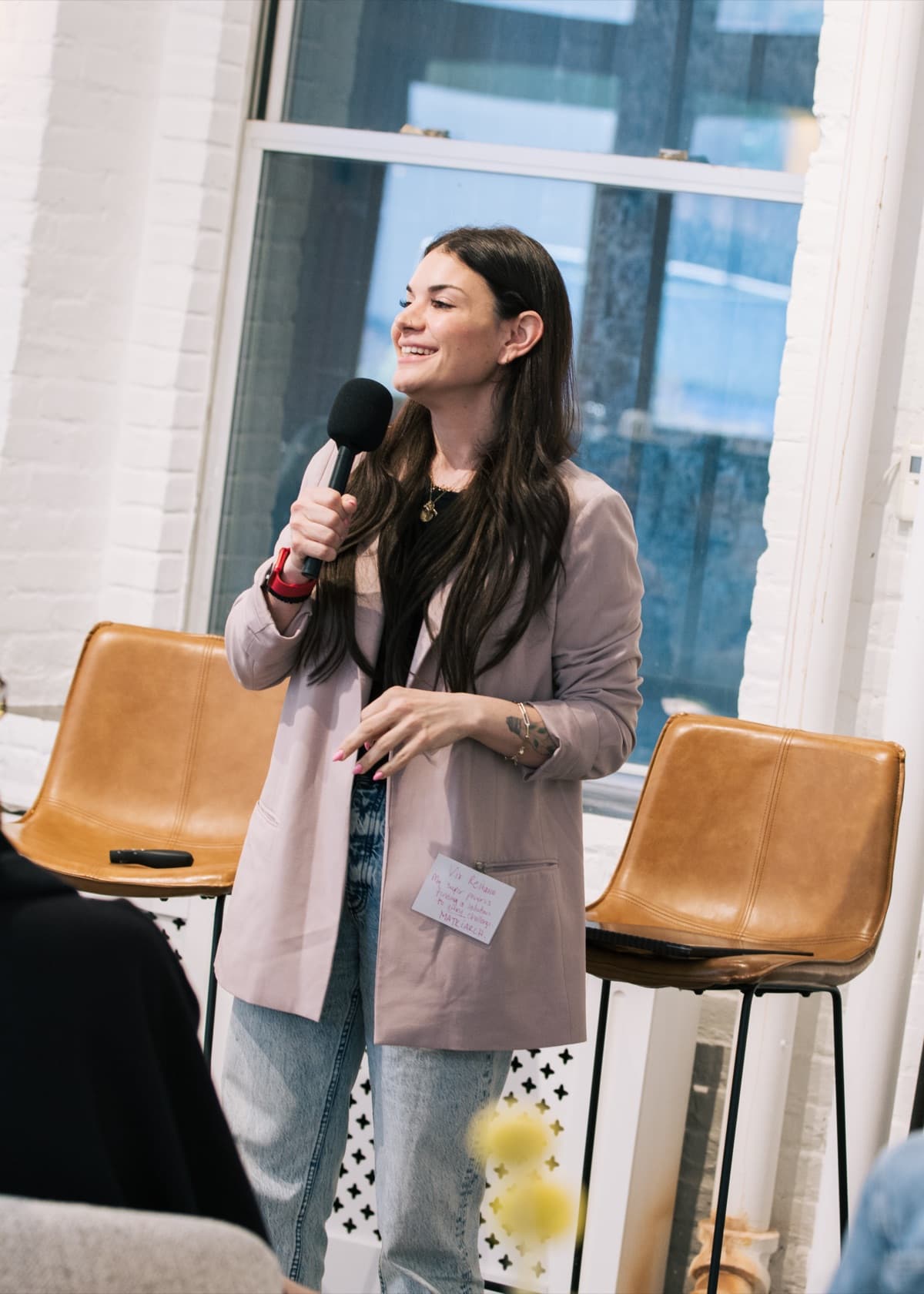 Vix Reitano speaking into a microphone at a live Marsha event, smiling, in a blush blazer in front of a white brick wall
