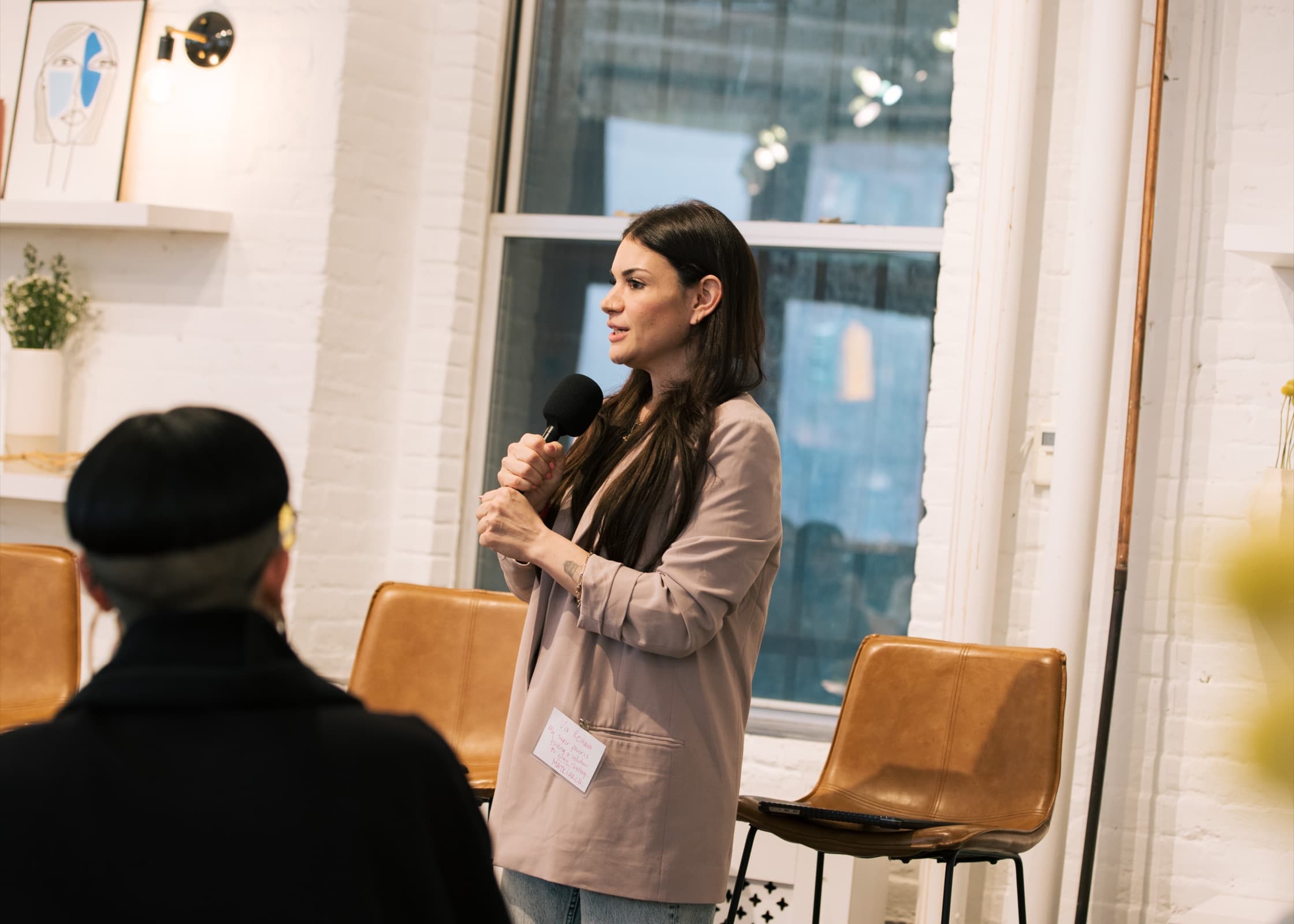 Vix Reitano holding a microphone and speaking to a seated audience at a Marsha event, in a blush blazer in front of a white brick wall